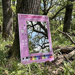 Può includere: Uno specchio con cornice rosa progettato per assomigliare alla finestra del programma "Paint" classico. Lo specchio presenta una barra degli strumenti con icone e testo, tra cui "File", "Edit" e "Colors". Il testo "untitled - Paint" è in alto. Lo specchio è appoggiato a un albero.