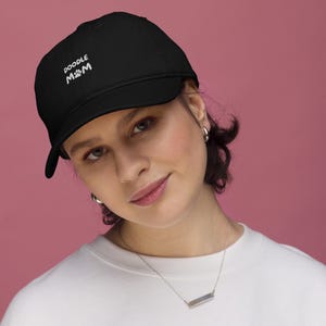 Puede incluir: Gorra de béisbol negra con el texto "Doodle Mom" en blanco, con una huella de pata. La gorra tiene una visera curva. El fondo es de color rosa liso.