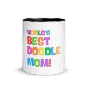 以下が含まれることがあります： 黒い内側の縁が付いた白いセラミックマグカップ。「WORLD'S BEST DOODLE MOM!」というテキストがカラフルな太字で表示されています。犬好きの方へのプレゼントに最適です。