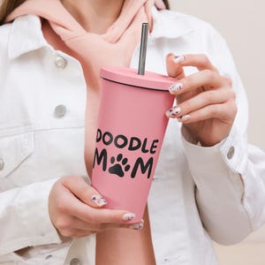 Peut inclure: Un gobelet rose avec un couvercle noir et une paille argentée. Le gobelet porte l'inscription "DOODLE MOM" avec un motif de patte. Le gobelet est tenu par une personne portant une veste blanche.