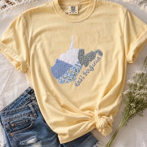 May include: A pale yellow short-sleeve t-shirt featuring a central applique of the state of West Virginia. The state shape is divided into sections of white, blue gingham, blue floral, and grey patterned fabric with the text West Virginia.