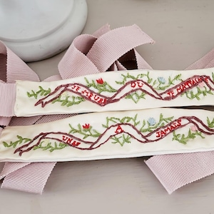 Può includere: Due nastri ricamati bianco sporco con testo bordeaux e accenti floreali. I nastri recitano "JE MEURS OU JE M'ATTACHE" e "UNAS JAMAIS". Nastro rosa a righe drappeggiato attorno ai nastri ricamati.