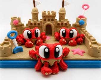 Puede incluir: Un castillo de arena impreso en 3D de color beige sobre una base azul, con tres figuras de cangrejos rojos con grandes ojos blancos y negros. La escena incluye una pala azul, una pala rosa, una estrella de mar rosa y conchas pequeñas.