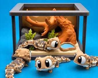 Puede incluir: Un recinto rectangular gris que contiene una figura de lagarto articulada de color tostado y marrón, un trozo de madera flotante marrón, rocas grises, plantas verdes y un pequeño cuenco de agua azul, con tres figuras de lagarto iguales delante.