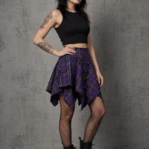 Op de afbeelding: Een zwarte crop top gecombineerd met een paarse geruite rok met een unieke asymmetrische zoom en decoratieve hardware. De rok heeft een zwart, wit en paars ruitpatroon. De outfit wordt gecompleteerd met zwarte laarzen en visnetkousen.