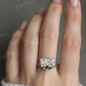 Op de afbeelding: Een zilverkleurige ring met twee rechthoekig geslepen heldere stenen die naast elkaar zijn geplaatst. De band vertoont ingewikkeld metaalwerk met een gestructureerd filigraanpatroon aan de zijkanten, wat het sieraad een gedetailleerd uiterlijk geeft.