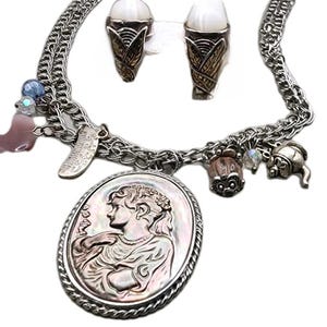 May include: A silver-coloured necklace and matching earrings set. The necklace has a large oval pendant with a woman's profile, and various charms. The earrings are shaped like ice cream cones. The set is on a white background.
