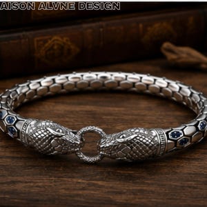 May include: A silver-toned metal bracelet featuring a textured, scale-like pattern along the band and two detailed snake head terminals that meet at a central circular ring. The design includes small blue accents embedded within the scales.