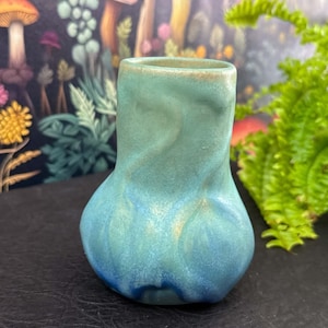 May include: A small, turquoise ceramic vase with a bulbous base and a narrow neck. The vase has a textured, organic shape and a gradient of colors from light green to deep blue. The background features a whimsical mushroom-themed wallpaper and green foliage.