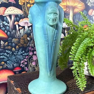 May include: A turquoise ceramic vase with a detailed relief of a Native American face. The vase has a flared base and a wide, open top. The face is intricately carved, with detailed features. The vase is a unique decorative item.