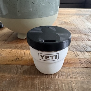 May include: A white YETI tumbler with a textured finish and a black lid. The YETI brand name is printed in a rectangular box on the front of the cup. The tumbler has a tapered shape and a wide opening at the top, designed for holding beverages.
