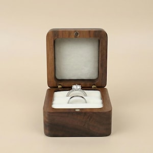 May include: A dark wooden ring box with a hinged lid, open to reveal a diamond engagement ring and a wedding band on a white cushion. The box is square with a smooth finish, set against a neutral background.