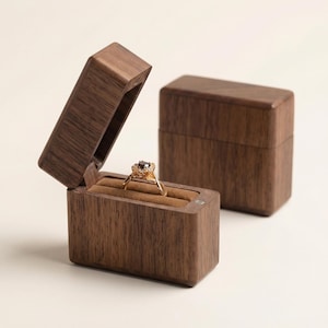 Puede incluir: Un joyero de madera marrón oscuro, uno abierto para revelar un anillo de oro con una piedra preciosa oscura. La caja tiene forma rectangular y una tapa con bisagras. Una segunda caja cerrada es visible en el fondo.