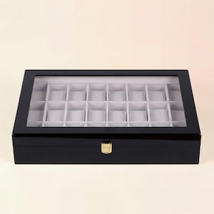 May include: A black, rectangular watch display case with a clear glass top. The interior is divided into multiple compartments with gray padded cushions. A gold-colored clasp secures the lid.