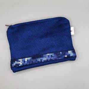 Peut inclure: Une pochette zippée bleu marine avec une bande de sequins bleus au bas. La pochette est rectangulaire et semble être faite d'une matière douce semblable à du daim. Une petite étiquette blanche est attachée sur le côté.