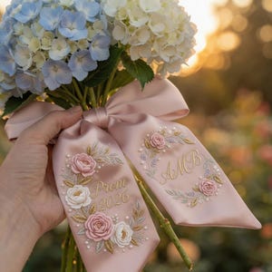Könnte beinhalten: Ein Strauß aus blauen und weißen Hortensien, gebunden mit einem hellrosa Satinband. Das Band zeigt gestickte Blumendesigns, Perlen und den Text "Prom 2026" und Initialen.
