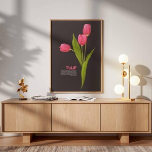 May include: A framed art print featuring three pink tulips with green leaves against a dark background. The word "TULIP" is displayed in pink. The print hangs above a wooden cabinet with a gold lamp and decorative items.