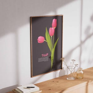 May include: A framed art print featuring three pink tulips with green leaves against a dark gray background. The word "TULIP" is displayed in pink. The print is mounted on a wall above a wooden surface.