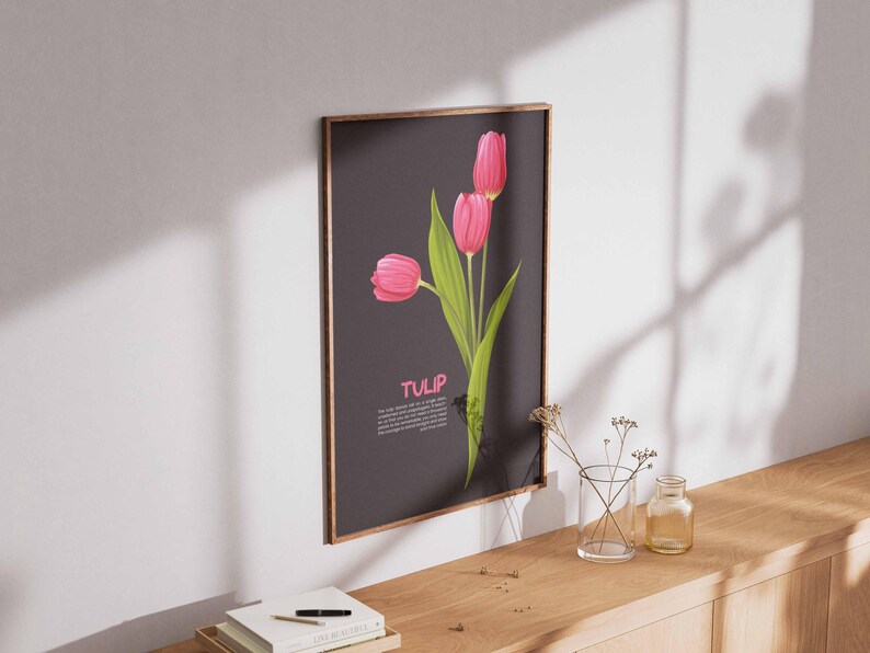 May include: A framed art print featuring three pink tulips with green leaves against a dark gray background. The word "TULIP" is displayed in pink. The print is mounted on a wall above a wooden surface.