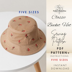 Könnte beinhalten: Ein beigefarbener Fischerhut mit roten Blumenstickereien. Der Hut liegt auf einem weißen Tisch. Auf der rechten Seite steht "Classic Bucket Hat Sewing Pattern" und "FIVE SIZES".