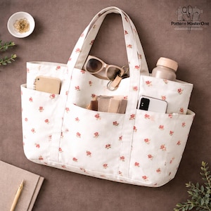 Pu&ograve; includere: Una borsa tote bianca con un motivo floreale di piccole rose rosse. La borsa ha pi&ugrave; tasche e contiene oggetti come occhiali da sole, un telefono e un taccuino. La borsa ha due manici.