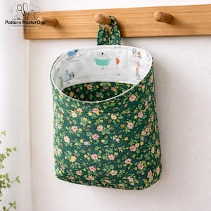 May include: A hanging fabric storage basket with a green floral pattern. The interior lining is white with cartoon illustrations. The basket is suspended from a wooden rack with two pegs.