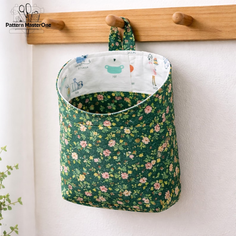 May include: A hanging fabric storage basket with a green floral pattern. The interior lining is white with cartoon illustrations. The basket is suspended from a wooden rack with two pegs.