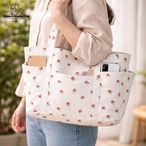 Pu&ograve; includere: Una borsa tote bianca con un motivo floreale di piccoli fiori rossi. La borsa ha pi&ugrave; tasche ed &egrave; portata da una persona. La borsa &egrave; realizzata in un tessuto robusto e ha un design semplice. La borsa ha un telefono e un taccuino nelle tasche.