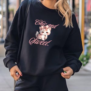 Puede incluir: Conjunto de sudadera y shorts negros. La sudadera presenta un gráfico de un gatito con un lazo rosa y el texto "Chic and Sweet" en escritura rosa. El conjunto es informal y cómodo.
