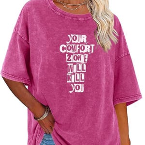 May include: A magenta-colored, short-sleeved t-shirt with the text "YOUR COMFORT ZONE WILL KILL YOU" in white block letters. The shirt has a slightly faded, vintage look.