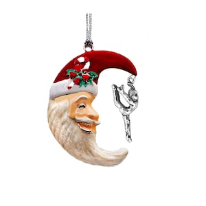 May include: A Christmas ornament featuring a crescent moon with a Santa Claus face, wearing a red hat with white trim and holly. A silver ballerina charm hangs from the ornament. The ornament is suspended by a silver chain.