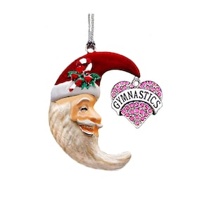 May include: A Christmas ornament featuring a crescent moon-shaped Santa Claus with a red hat and white beard. A heart-shaped charm with the word "GYMNASTICS" in white letters is attached. The charm is decorated with pink rhinestones.