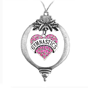 May include: Silver-toned ornament with a heart-shaped charm. The heart is filled with pink rhinestones and features the word "GYMNASTICS" in a white banner. The ornament has a decorative top with holly leaves and a loop for hanging.
