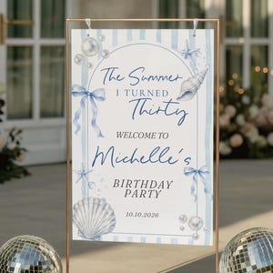Puede incluir: Un letrero rectangular para fiesta de cumpleaños en un soporte de metal color cobre, con rayas verticales azul claro, ilustraciones de conchas, estrellas de mar, perlas y cintas azules, con el texto The Summer I Turned Thirty Welcome to Michelle's Birthday Party 10.10.2026.