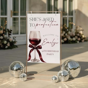 May include: A sign with the text "She's Aged to Perfection" and "Welcome to Emily's 30th Birthday Party" features a wine glass illustration. The sign is in a copper-colored frame, with disco balls at the base.