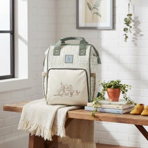 May include: A light green and white floral print nappy bag with a front pocket featuring an embroidered Winnie the Pooh design. The bag has a top handle and is on a wooden bench with a blanket and books.