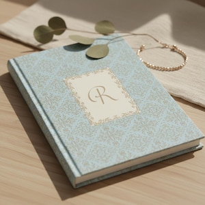 Puede incluir: Un diario azul claro con un patrón floral y la letra "R" en un rectángulo crema. Una pulsera dorada y hojas decorativas están colocadas cerca del libro cerrado. Ideal para escribir.