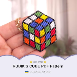 May include: A colorful, beaded Rubik's Cube pendant with a gold chain. The cube features red, orange, yellow, green, blue, and white squares. The image includes the text "RUBIK'S CUBE PDF Pattern" and "Design by Anastasiia Bozinian."