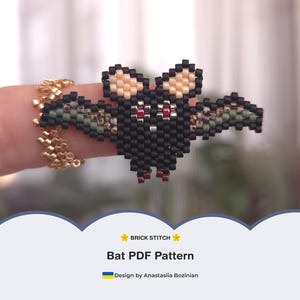 May include: A beaded bat ring in black, tan, red, and green. The bat is made with small beads and has outstretched wings. The ring band is gold-colored. The image also includes the text "BRICK STITCH" and "Bat PDF Pattern".