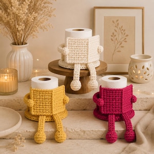 May include: Three toilet paper roll covers crocheted in cream, mustard yellow, and magenta. Each cover features a rectangular book-shaped front, two small arms, and two dangling legs with round feet, designed to hold a standard toilet paper roll.