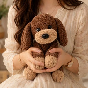 May include: A handmade brown and tan crochet dog plushie with large floppy ears and a black nose. The dog has tan paws and a tan snout. The plush toy is being held in someone's hands.