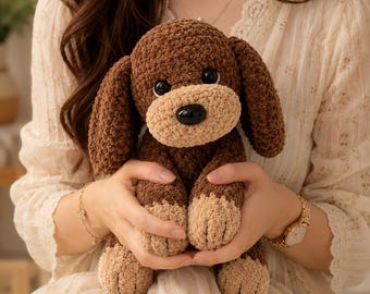 Puede incluir: Un peluche de perro de ganchillo marrón y beige hecho a mano con grandes orejas caídas y una nariz negra. El perro tiene patas beige y un hocico beige. El peluche está siendo sostenido en las manos de alguien.