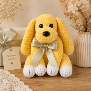 May include: A yellow crocheted dog plush toy with long floppy ears, white paws, and a black plastic nose. The dog features a smooth light green satin ribbon tied in a bow around its neck. The toy has round black eyes and a soft yarn texture.