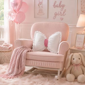 Peut inclure: Un fauteuil à bascule rembourré rose clair avec un coussin blanc en forme de nœud aux bords volantés et une bande centrale rose. Un plaid en tricot rose texturé est posé sur l'accoudoir et une peluche lapin crème est posée à côté.