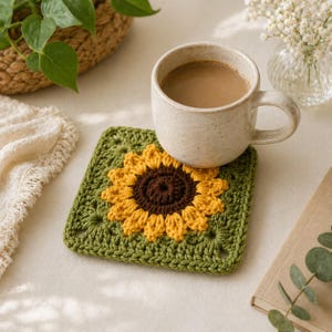 May include: A beige ceramic mug filled with coffee sits on a handmade crochet coaster. The coaster features a sunflower design with yellow petals and a brown center, set against a green background. A woven basket and a book are in the background.