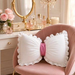 Könnte beinhalten: Ein weißes, schleifenförmiges Dekokissen mit gerüschten Kanten und einem einfarbigen rosa Mittelband. Das Kissen liegt auf einem rosa Samtstuhl und besteht aus weichem Stoff mit einem gerafften Design, das wie eine dekorative Schleife geformt ist.