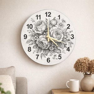 May include: Circular wall clock with a floral design. The clock face has a black and white illustration of roses and other flowers. Black numerals mark the hours, and the hands are gold. The clock is mounted on a white wall.