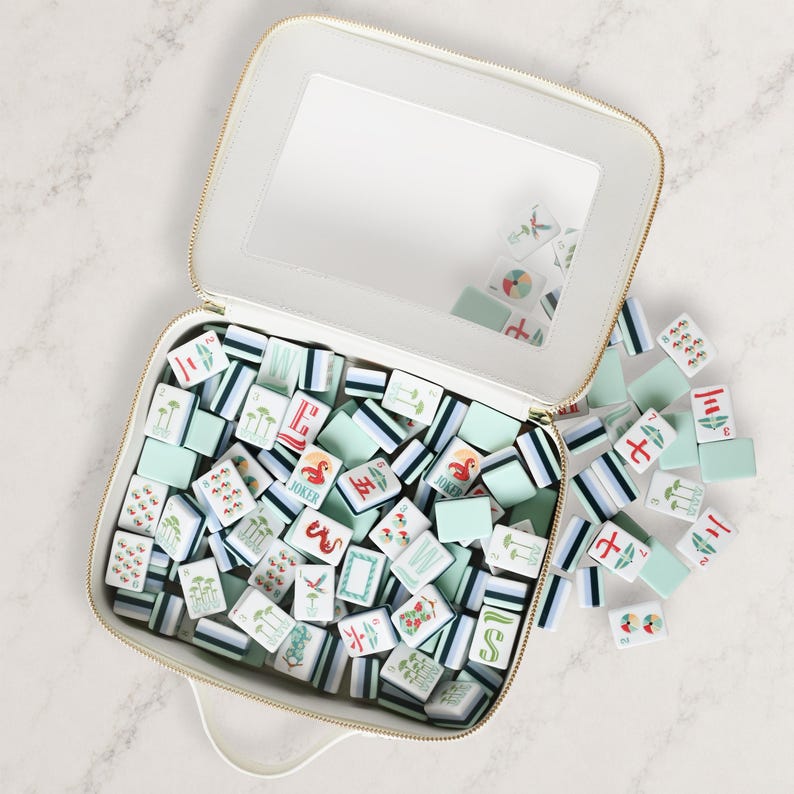 May include: A white zippered carrying case with a clear window lid, filled with a collection of rectangular mahjong tiles. The tiles feature various colorful illustrations, including palm trees, flamingos, and Chinese characters in pastel tones.