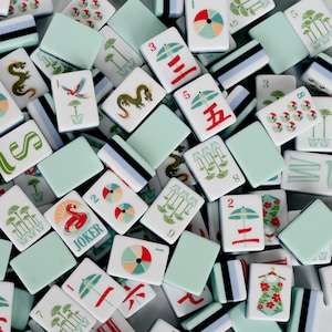 May include: A collection of rectangular mahjong tiles featuring a mix of solid light green backs and white faces with colorful illustrations. Designs include red umbrellas, green dragons, palm trees, a flamingo labeled JOKER, and floral swimsuits.
