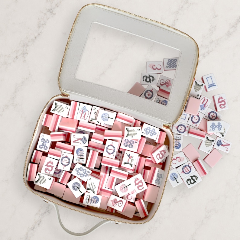 May include: A rectangular white carrying case with a clear window lid, filled with a set of mahjong tiles. The tiles feature pink, blue, and white color schemes with various illustrations, numbers, and the text JOKER printed on some pieces.